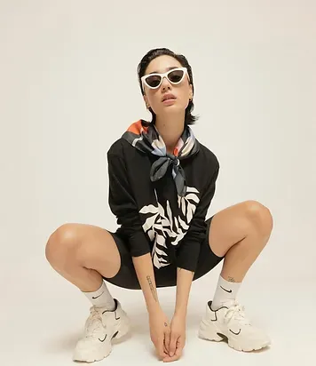 Modelo agachada con lentes de sol blancos, pañuelo estampado y polerón negro