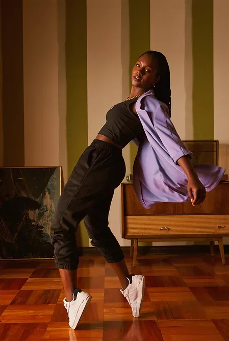 Modelo con chaqueta lila y pantalón negro posando en una sala de estar retro