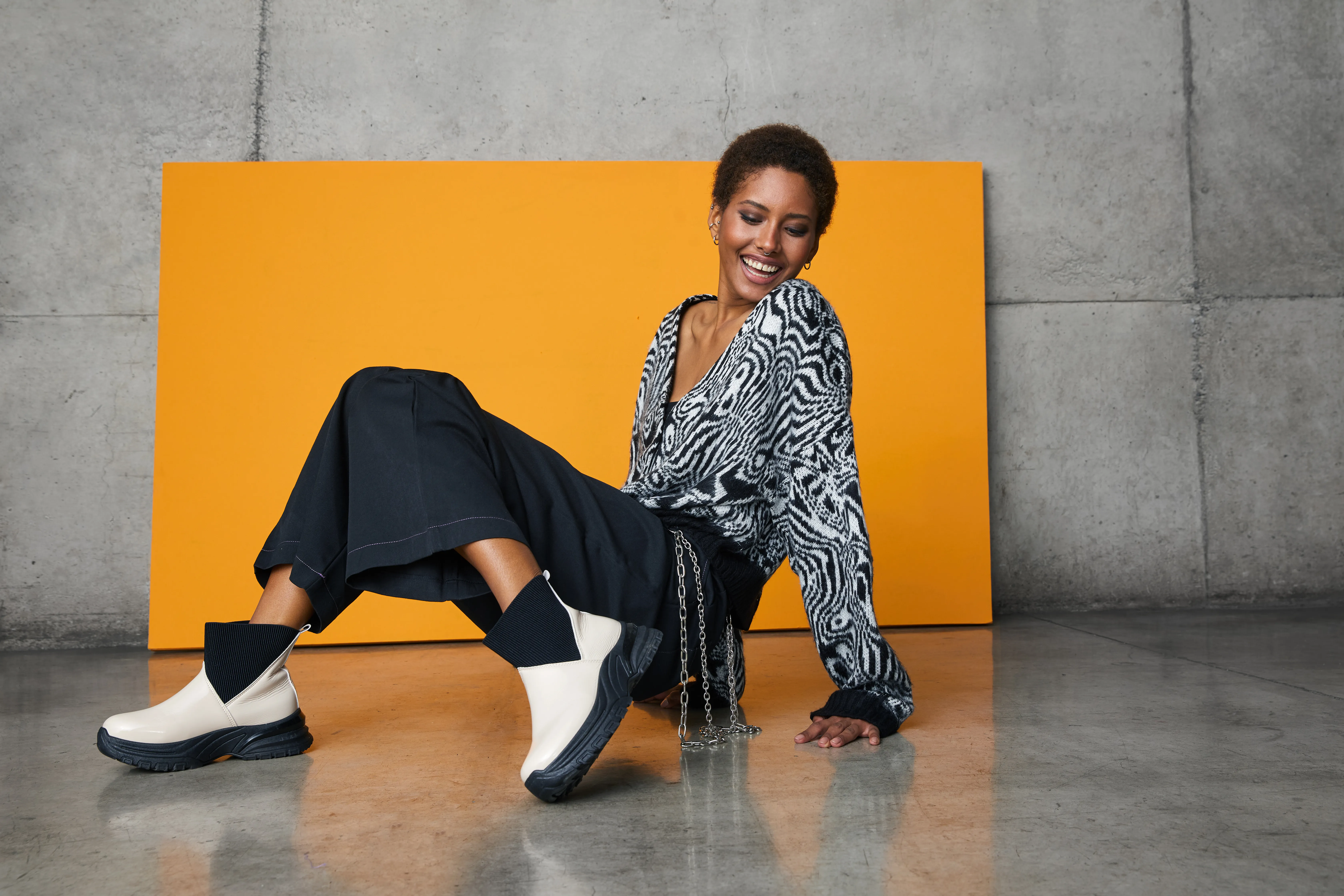 Modelo sentada con blusa estampada, pantalón negro y zapatillas blancas frente a un panel naranjo