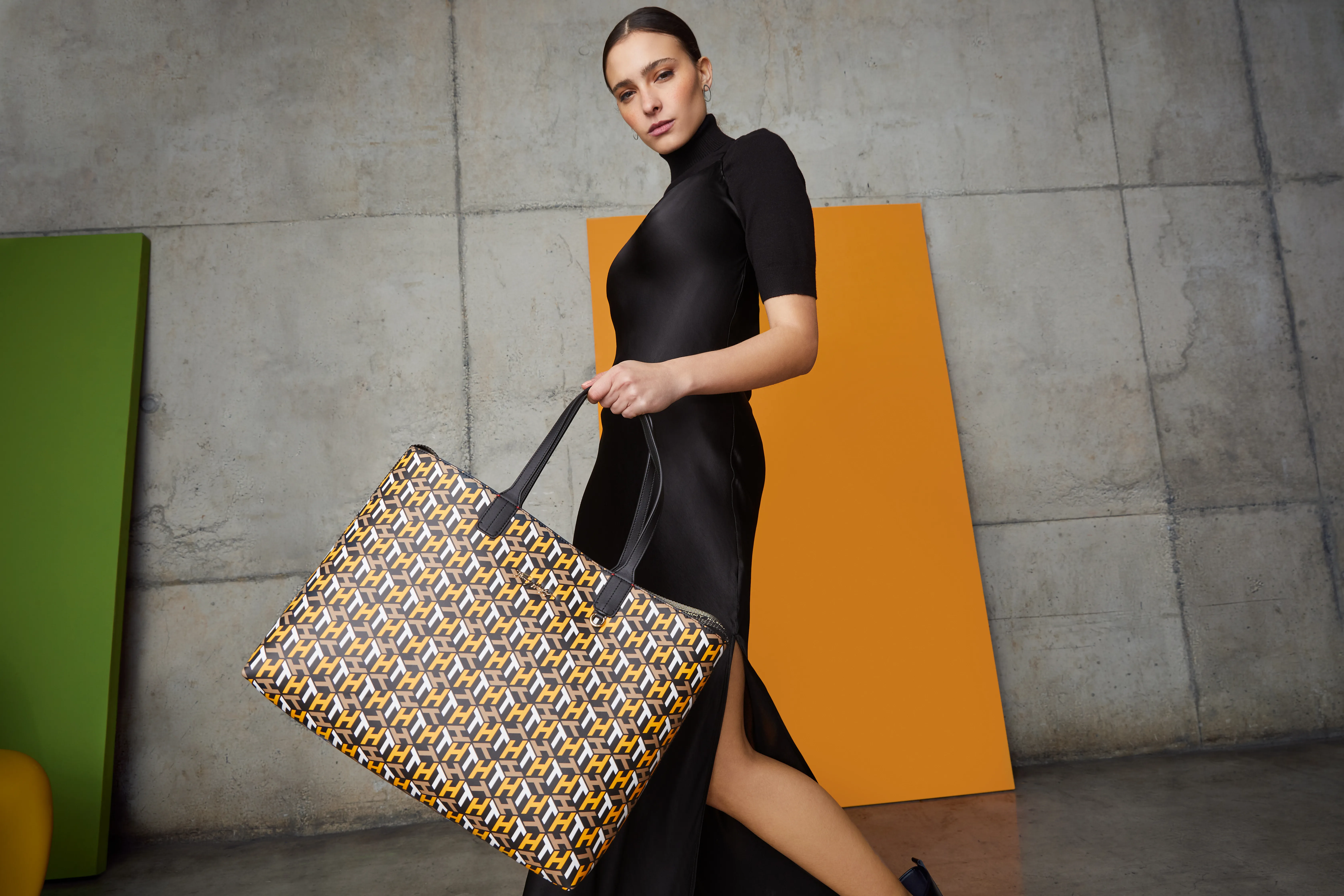 Modelo con vestido negro y cartera estampada frente a un panel naranjo